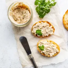 Pyszna pasta z łososia na chrupiących grzankach, ozdobiona natką pietruszki. Obok słoiczek z pastą i nóż.