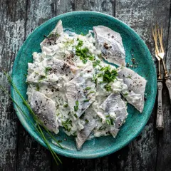 Śledzie w śmietanie, przepis babci, podane na turkusowym talerzu, posypane szczypiorkiem i pieprzem.