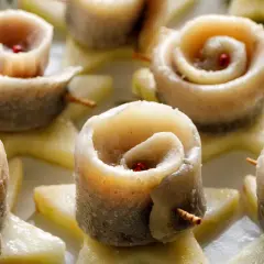 Roladki ze śledzi w occie, ozdobione ziarnami pieprzu, ułożone na plasterkach jabłka.