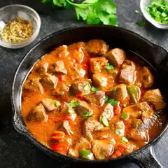 Gulasz z kurczaka w aromatycznym sosie, podany w żeliwnej patelni, z papryczkami chili i natką pietruszki.