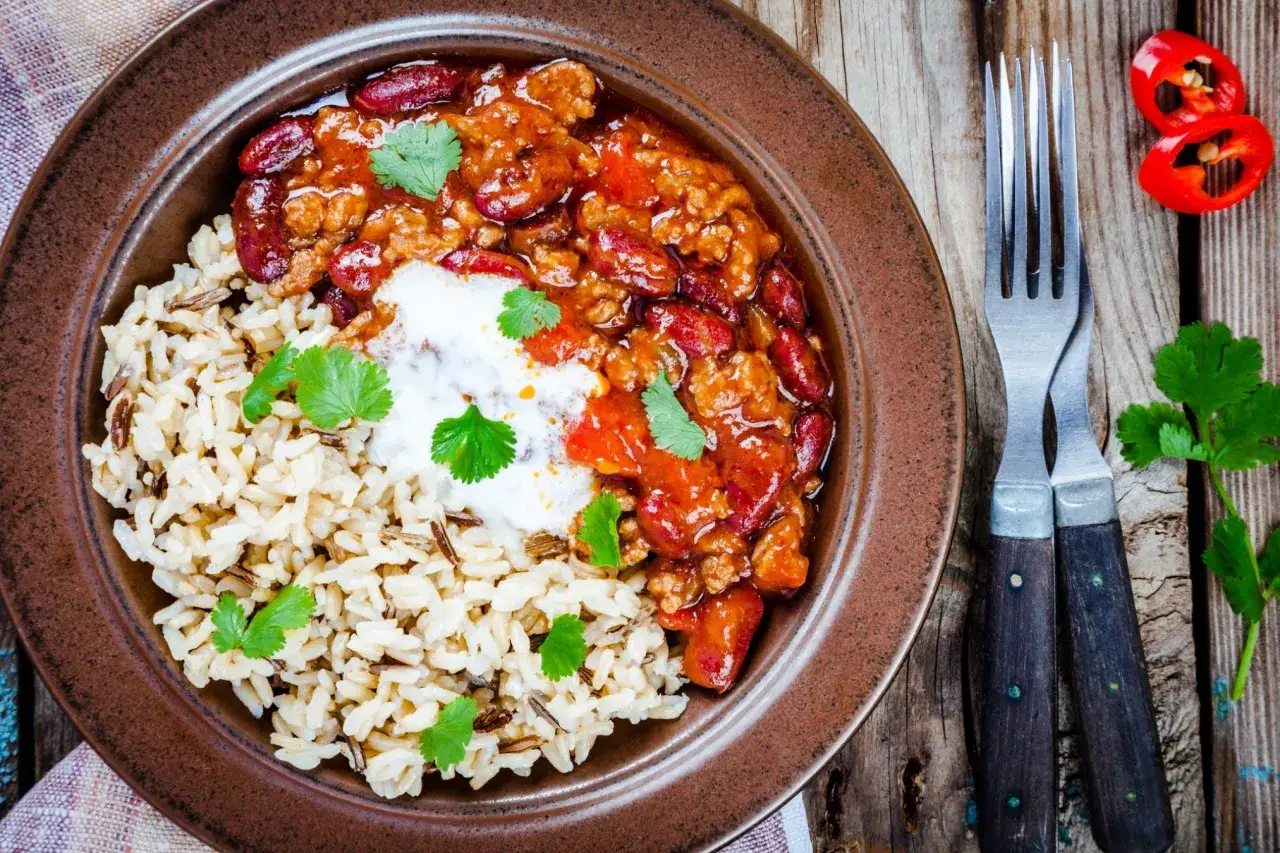 Pyszne chili con carne z ryżem, ozdobione kolendrą i śmietaną. Obok widelce i papryczki chili.