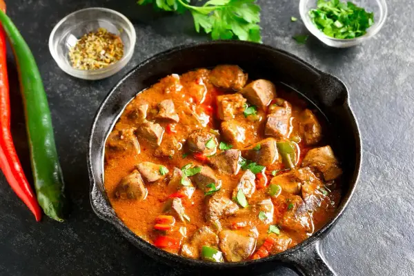 Gulasz z kurczaka w aromatycznym sosie, podany w żeliwnej patelni, z papryczkami chili i natką pietruszki.